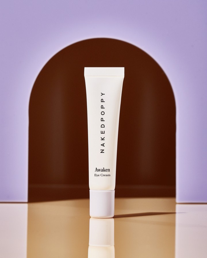 Product shot of Naked Poppy Awaken Eye Cream tube against lavender and brown archway background. Studio product photography for skincare.