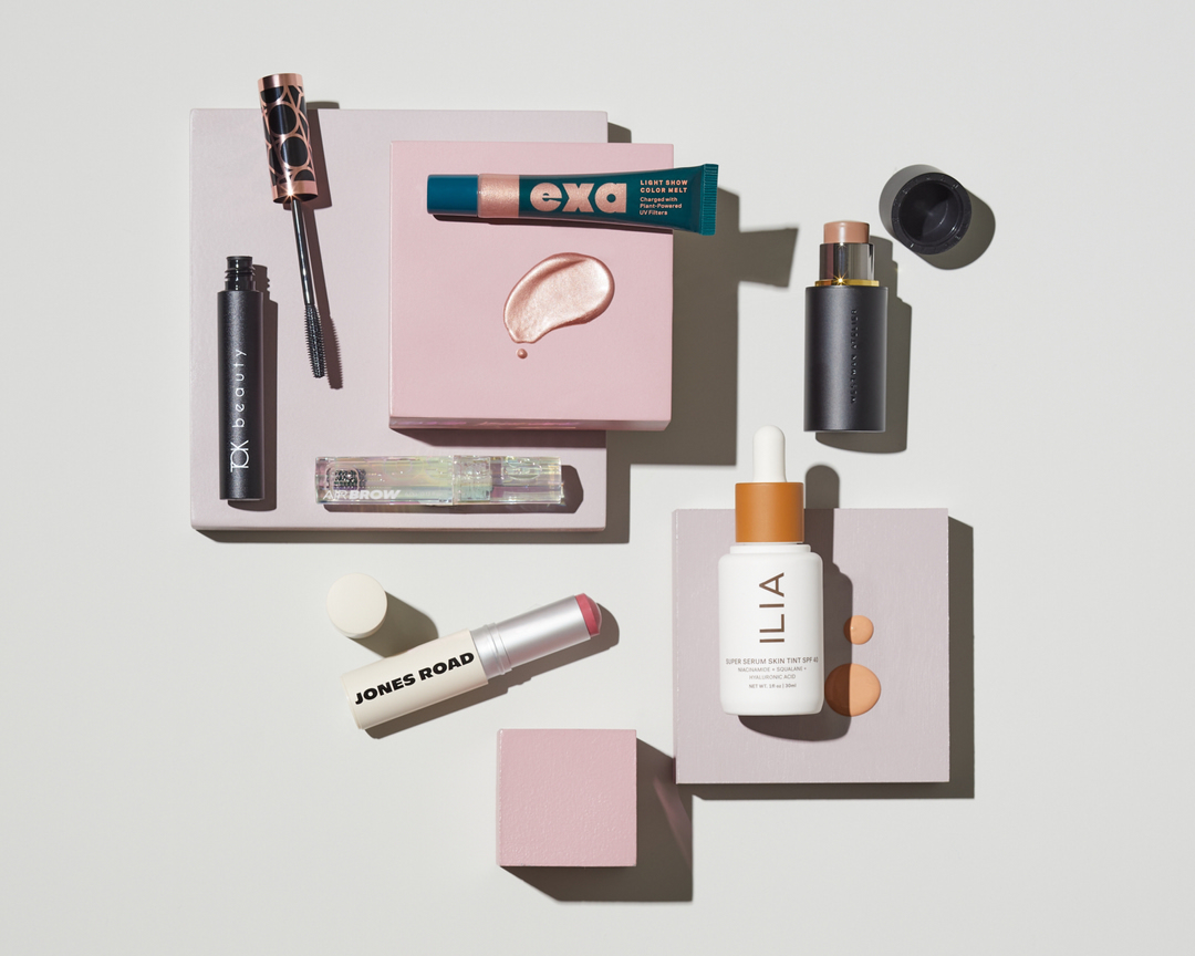Cosmetic products including mascara, eye serum, and lip balm are arranged on a pale pink and grey background in a beauty product flat lay.