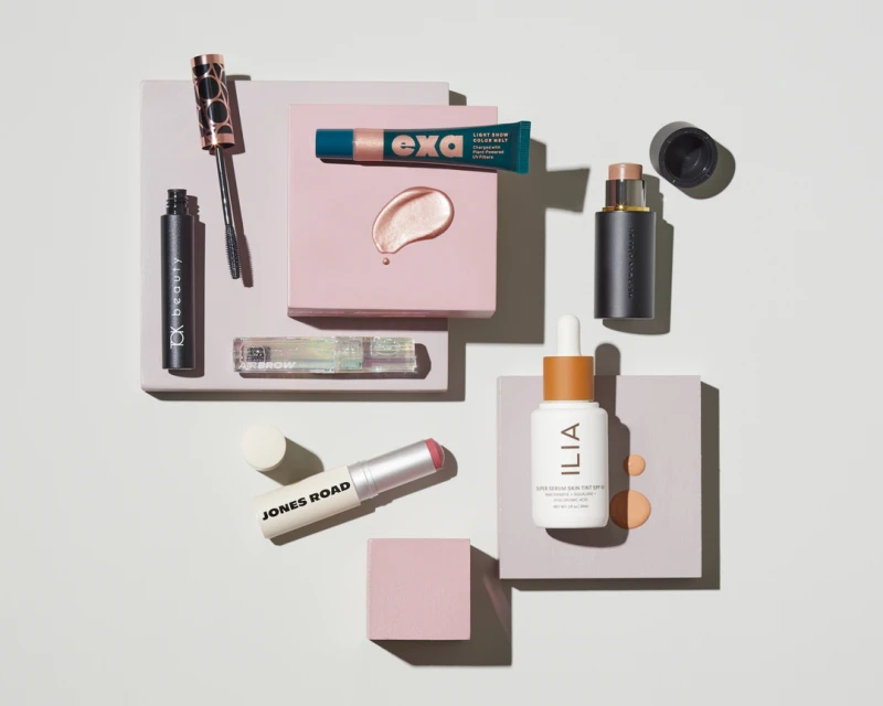 Cosmetic products including mascara, eye serum, and lip balm are arranged on a pale pink and grey background in a beauty product flat lay.