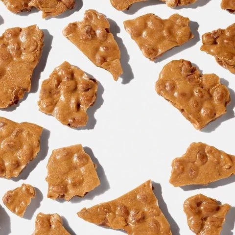 Overhead shot of peanut brittle pieces with light tan and golden brown colors, scattered on a plain white surface in a flat lay.