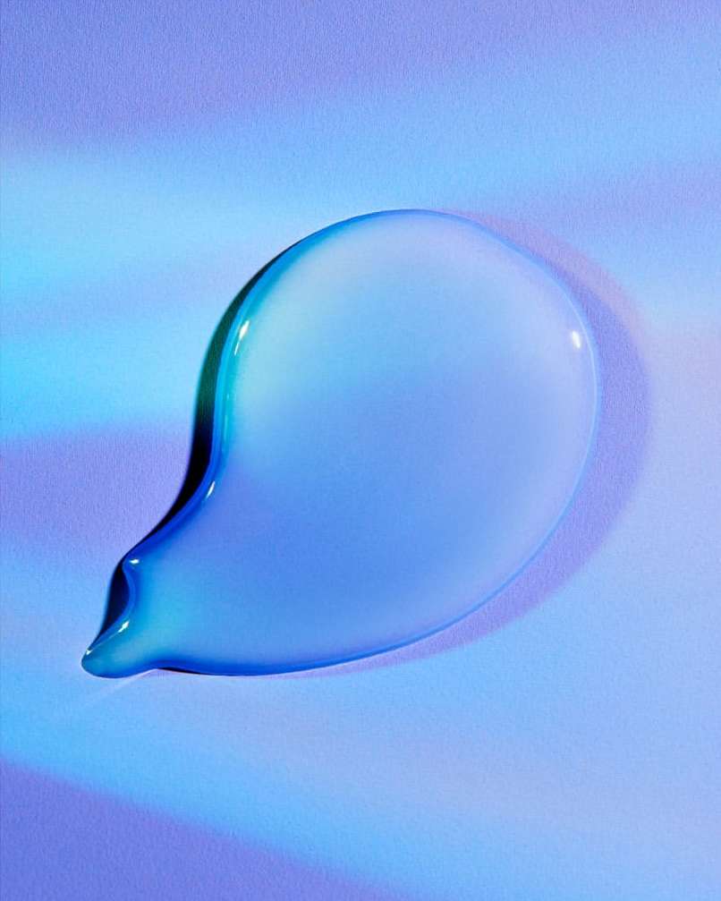 Close-up studio shot of a teardrop-shaped cosmetic liquid smear with a glossy, reflective surface over a gradient blue background.