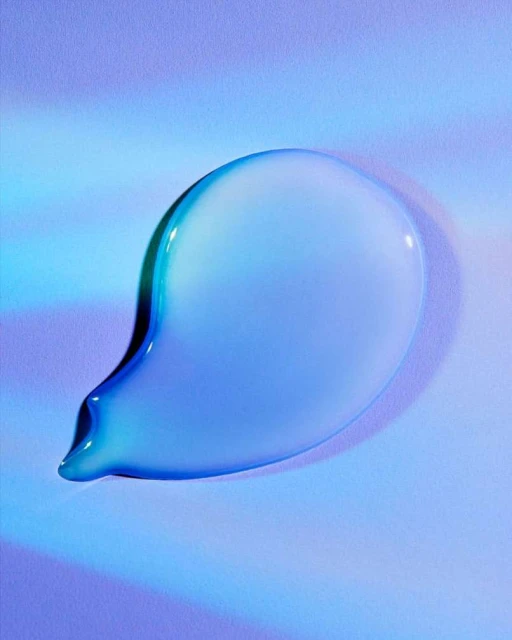 Close-up studio shot of a teardrop-shaped cosmetic liquid smear with a glossy, reflective surface over a gradient blue background.