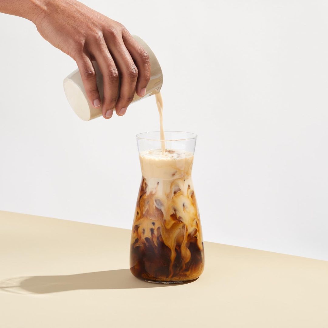 Iced coffee is poured from a glass jug into a glass carafe. The drink is on a cream-colored table with a white backdrop.