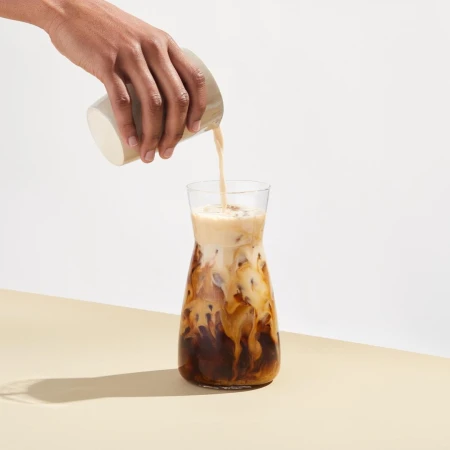 Iced coffee is poured from a glass jug into a glass carafe. The drink is on a cream-colored table with a white backdrop.