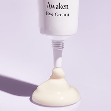 Close-up of white cream from a tube of 'Awaken Eye Cream' against a light purple background, highlighting the product's texture.