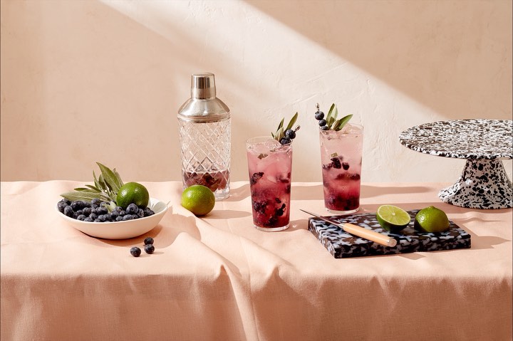 Two blueberry cocktails with limes and a cocktail shaker, on a table covered in a peach-colored linen cloth in a studio setting.