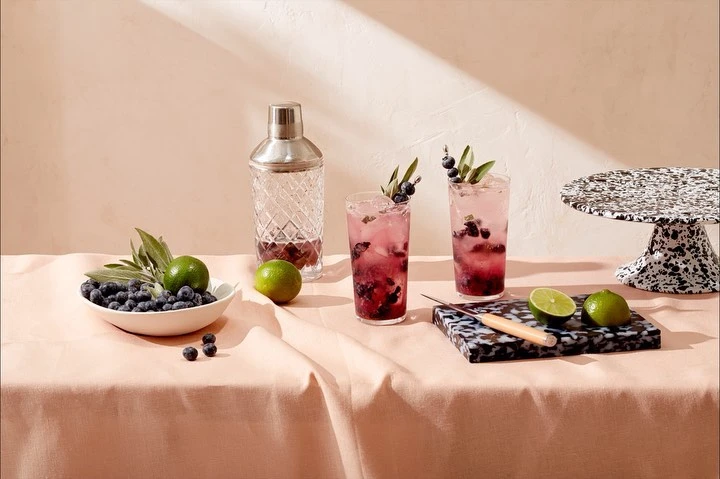 Two blueberry cocktails with limes and a cocktail shaker, on a table covered in a peach-colored linen cloth in a studio setting.