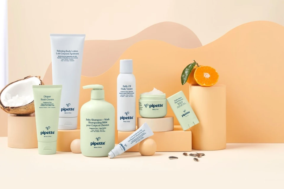 Collection of Pipette brand baby skincare products including lotion, cream, and shampoo, arranged on a geometric display with fruit and nuts.