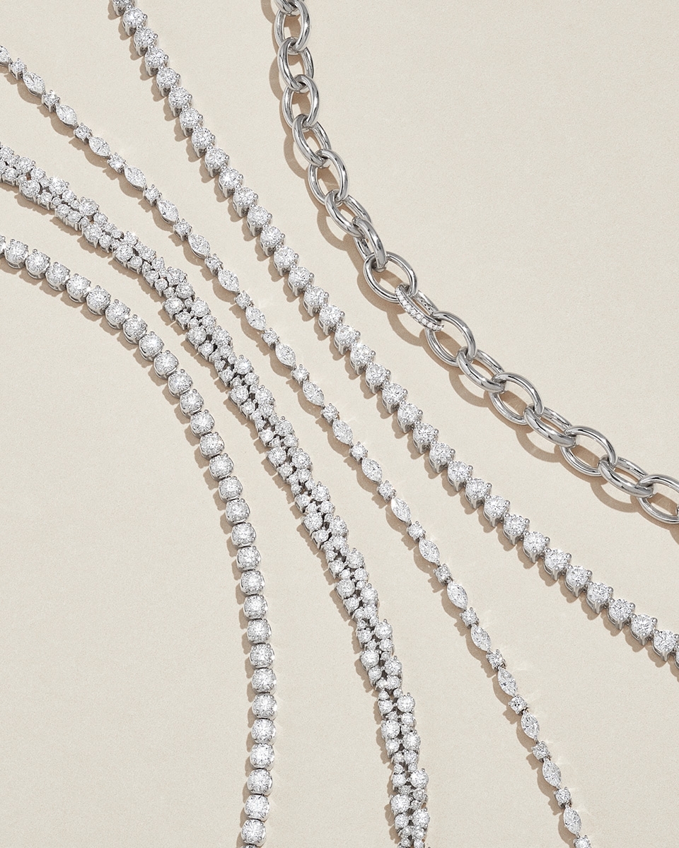 Overhead shot of an arrangement of diamond tennis bracelets and silver chain link necklaces on a beige background, showcasing jewelry.