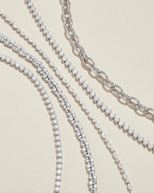 Overhead shot of an arrangement of diamond tennis bracelets and silver chain link necklaces on a beige background, showcasing jewelry.