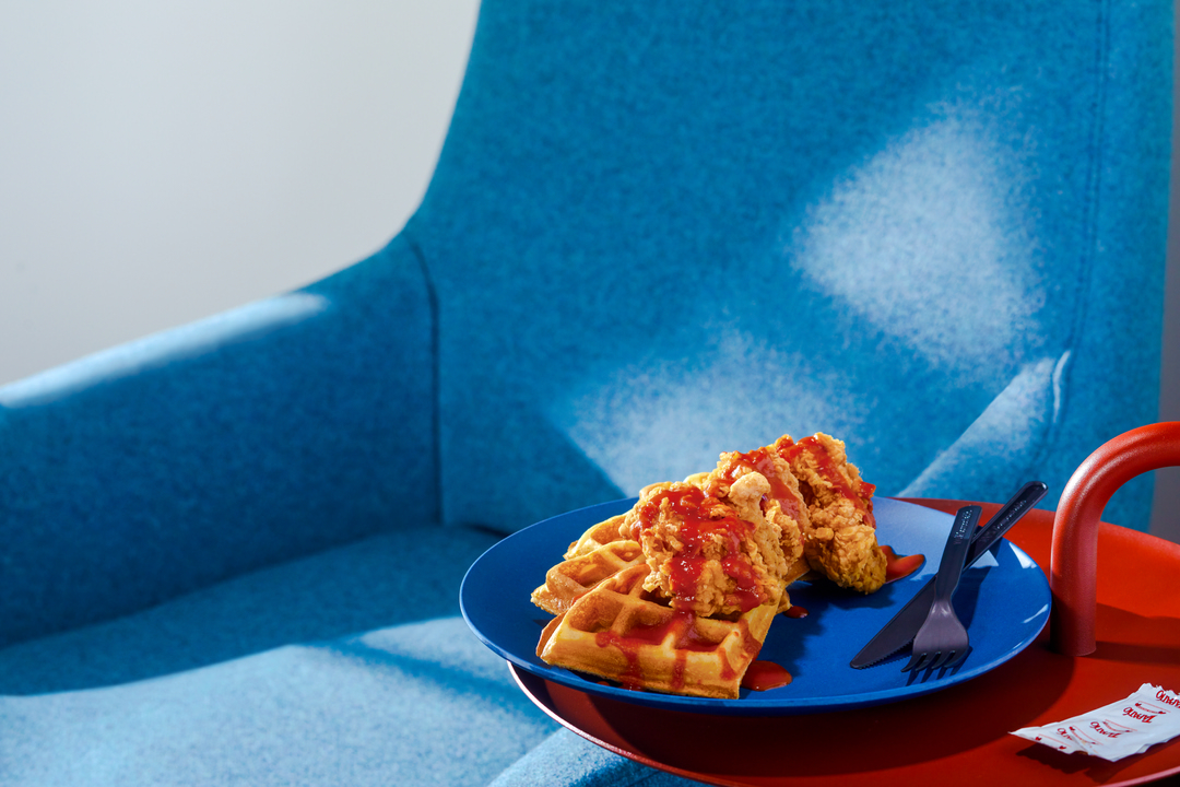 Golden waffles topped with fried chicken and a red sauce, plated on a blue plate alongside black cutlery on a red surface.