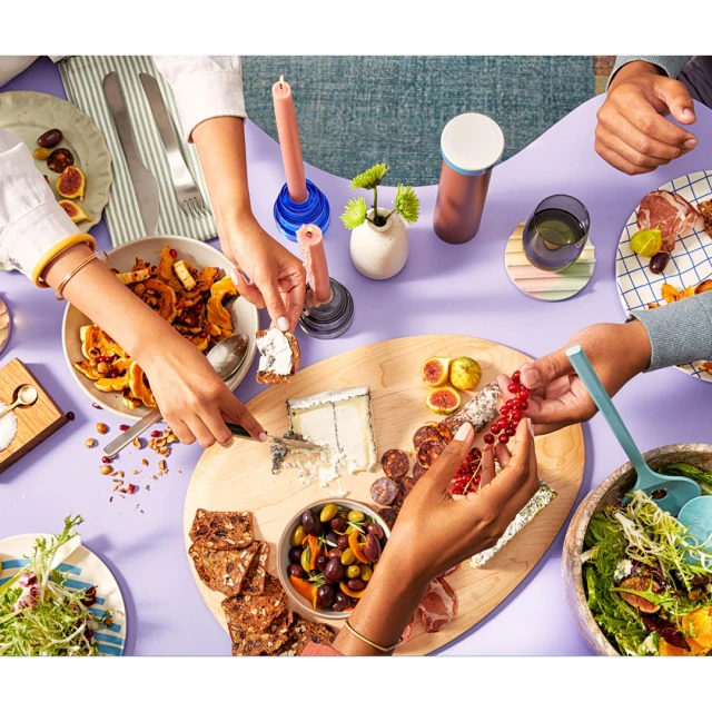 Overhead shot of a group sharing an appetizer spread on a purple table, featuring cheese, olives, fruit, and salads, with colorful tableware.