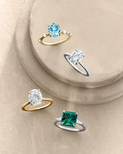 Four rings with varying gemstones on a multi-tiered beige surface, showcasing jewelry design and gem settings for an elegant presentation.