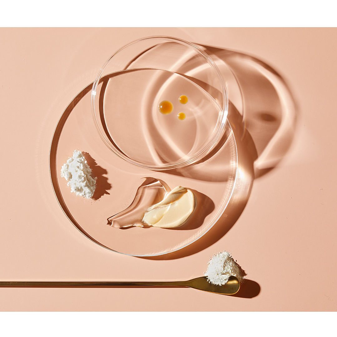 Beauty product ingredients arranged in petri dishes and on a golden spoon against a peach-toned backdrop, showcasing cosmetic science.