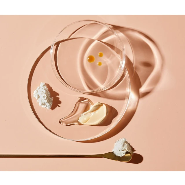 Beauty product ingredients arranged in petri dishes and on a golden spoon against a peach-toned backdrop, showcasing cosmetic science.