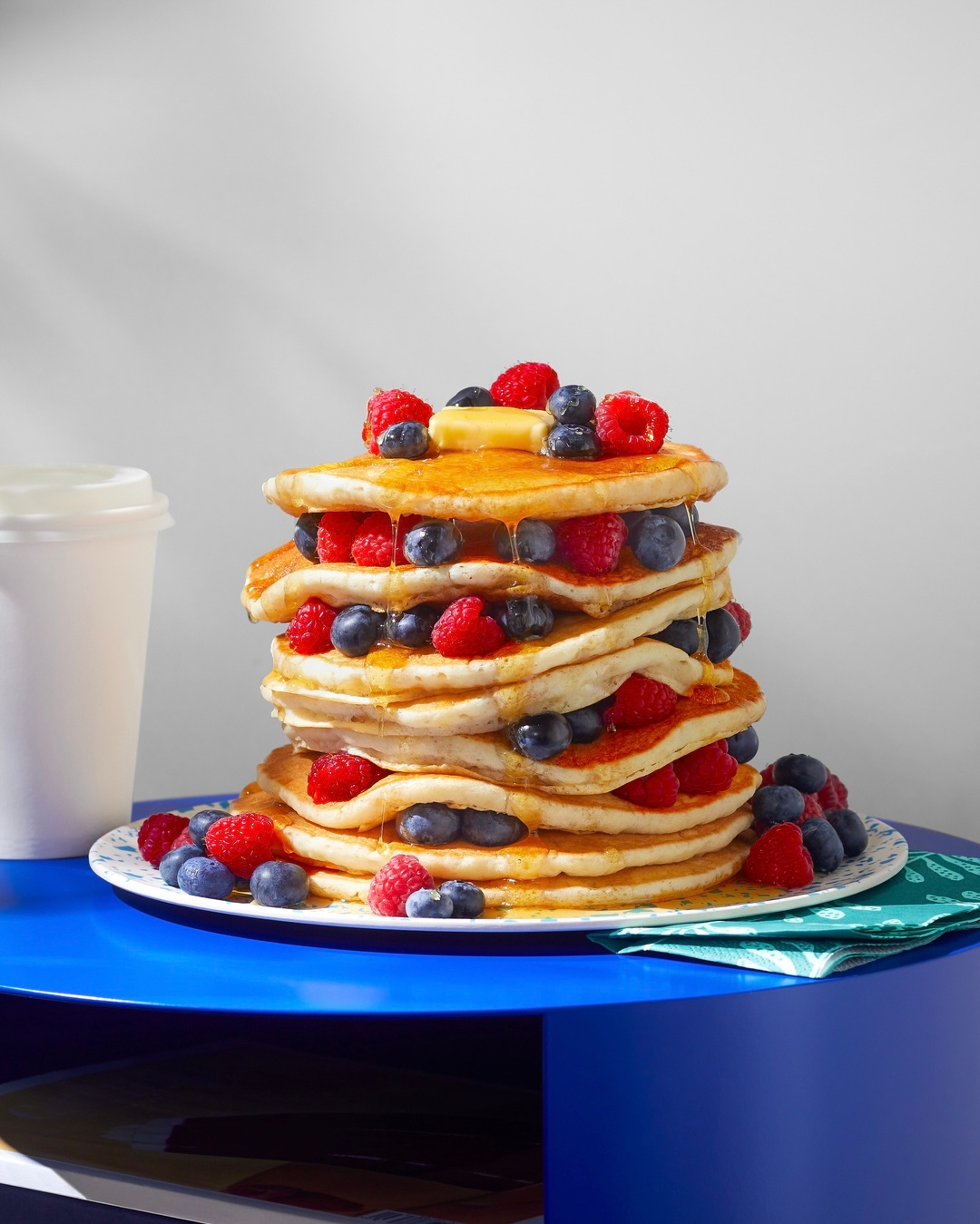 A tall stack of pancakes topped with fresh raspberries, blueberries, and a pat of butter, drizzled with syrup, on a blue table.
