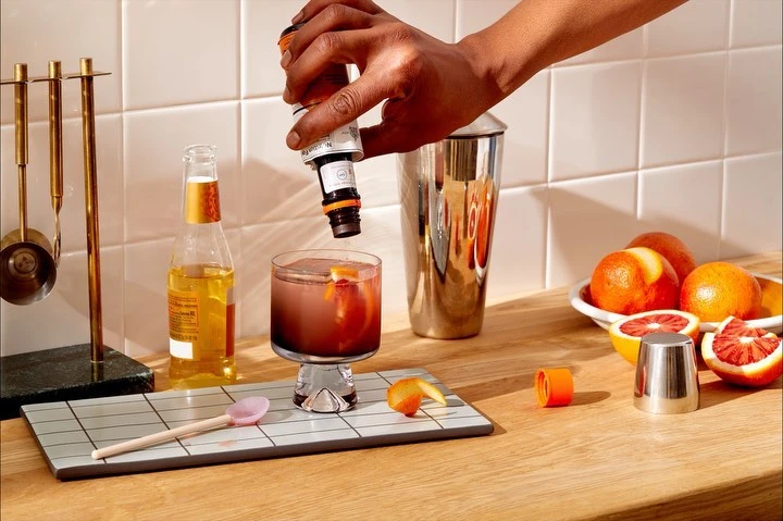 Cocktail preparation scene: Hand pouring bitters into a glass with ice and orange peel next to a shaker and a bottle of beer.