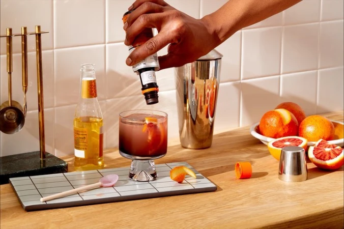 Cocktail preparation scene: Hand pouring bitters into a glass with ice and orange peel next to a shaker and a bottle of beer.