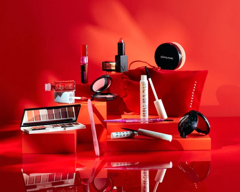 Still life of makeup products including lipstick, blush, mascara, eye shadow palette, and a red cosmetic bag on red background.