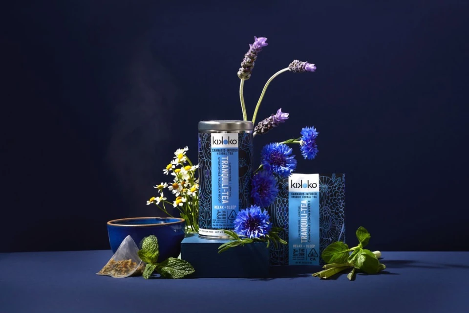 Still life of KIKOKO cannabis-infused herbal tea products including a tin, packet, tea bag, blue bowl, mint, chamomile, and lavender flowers.