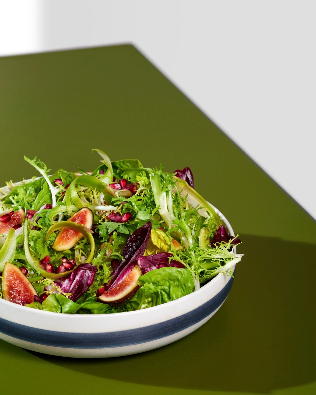 Fresh salad with mixed greens, figs, and pomegranate seeds in a white bowl, set on an olive-green surface against a white background.