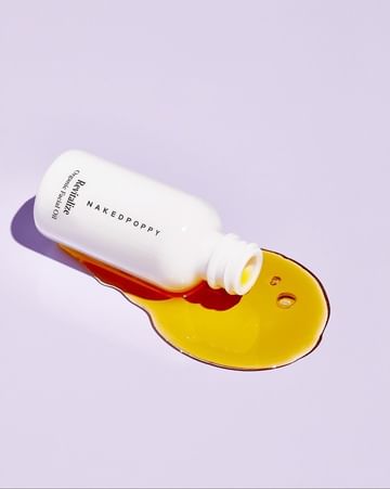 Bottle of revitalizing facial oil from Naked Poppy spills onto a light purple surface, showcasing its golden hue and minimalist design.