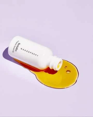 Bottle of revitalizing facial oil from Naked Poppy spills onto a light purple surface, showcasing its golden hue and minimalist design.