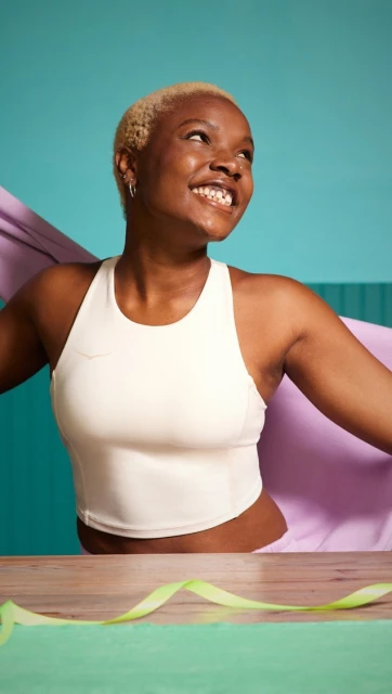 A smiling adult woman with short blonde hair wears a white crop top and poses indoors with a lavender fabric against a turquoise background.