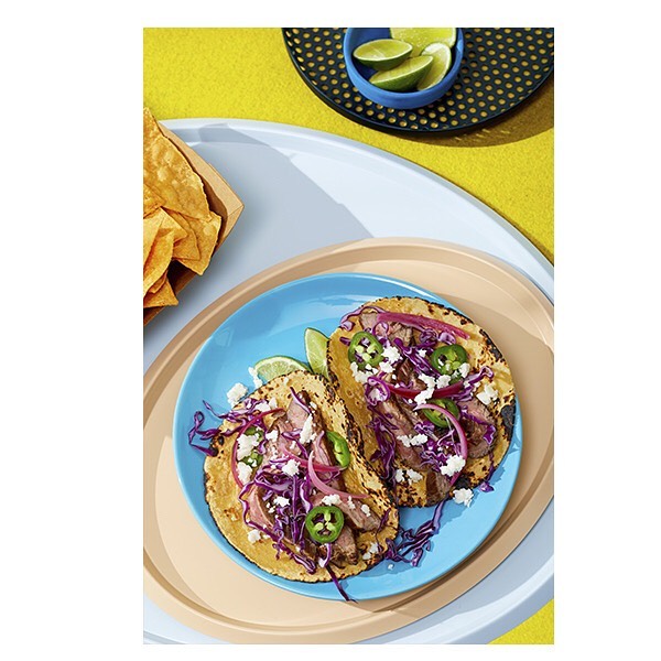 Overhead shot of two tacos with steak, cabbage, jalapeños, and cotija cheese on a blue plate, alongside lime wedges and chips.