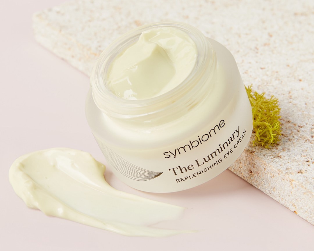 Eye cream jar with light yellow cream and a smear of cream on a pink surface with a textured stone slab in the background.