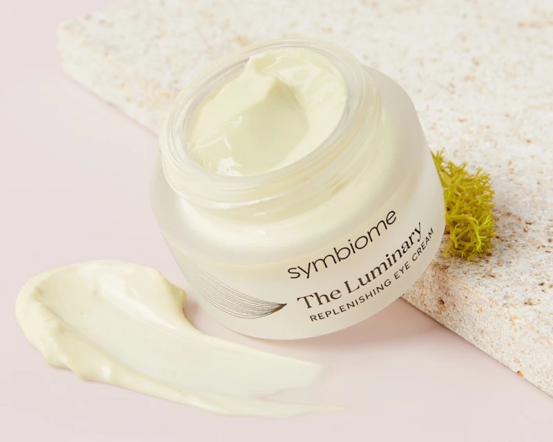 Eye cream jar with light yellow cream and a smear of cream on a pink surface with a textured stone slab in the background.