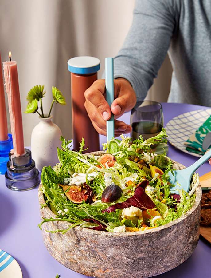 Overhead shot of a stone bowl filled with a fresh salad, set on a lilac table with candles, flowers, and a glass of water.