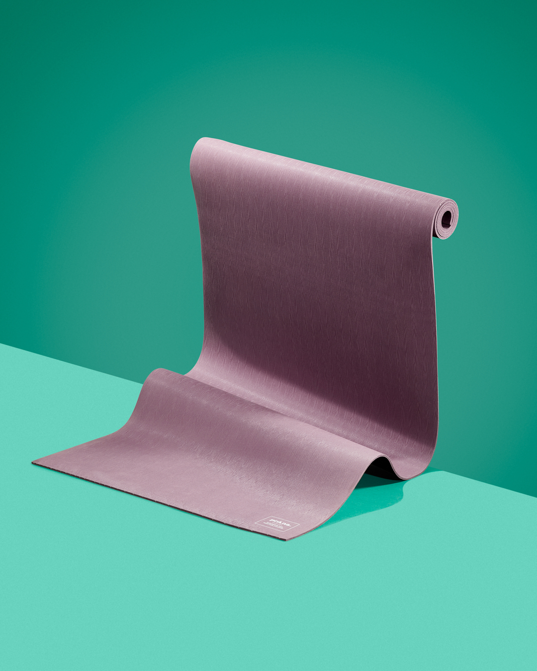 Unrolled yoga mat in light purple color on a two-tone background, displayed indoors in a studio environment with soft lighting.