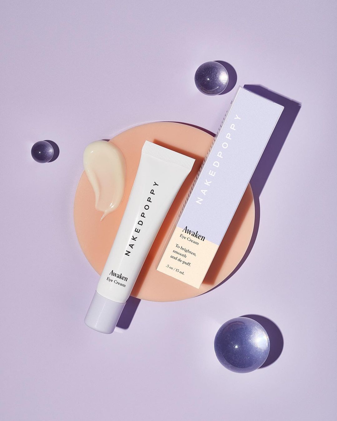 Overhead shot of NakedPoppy Awaken Eye Cream tube and box on a peach circle, surrounded by translucent spheres on a purple background.