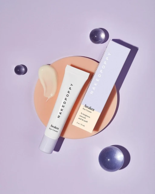 Overhead shot of NakedPoppy Awaken Eye Cream tube and box on a peach circle, surrounded by translucent spheres on a purple background.