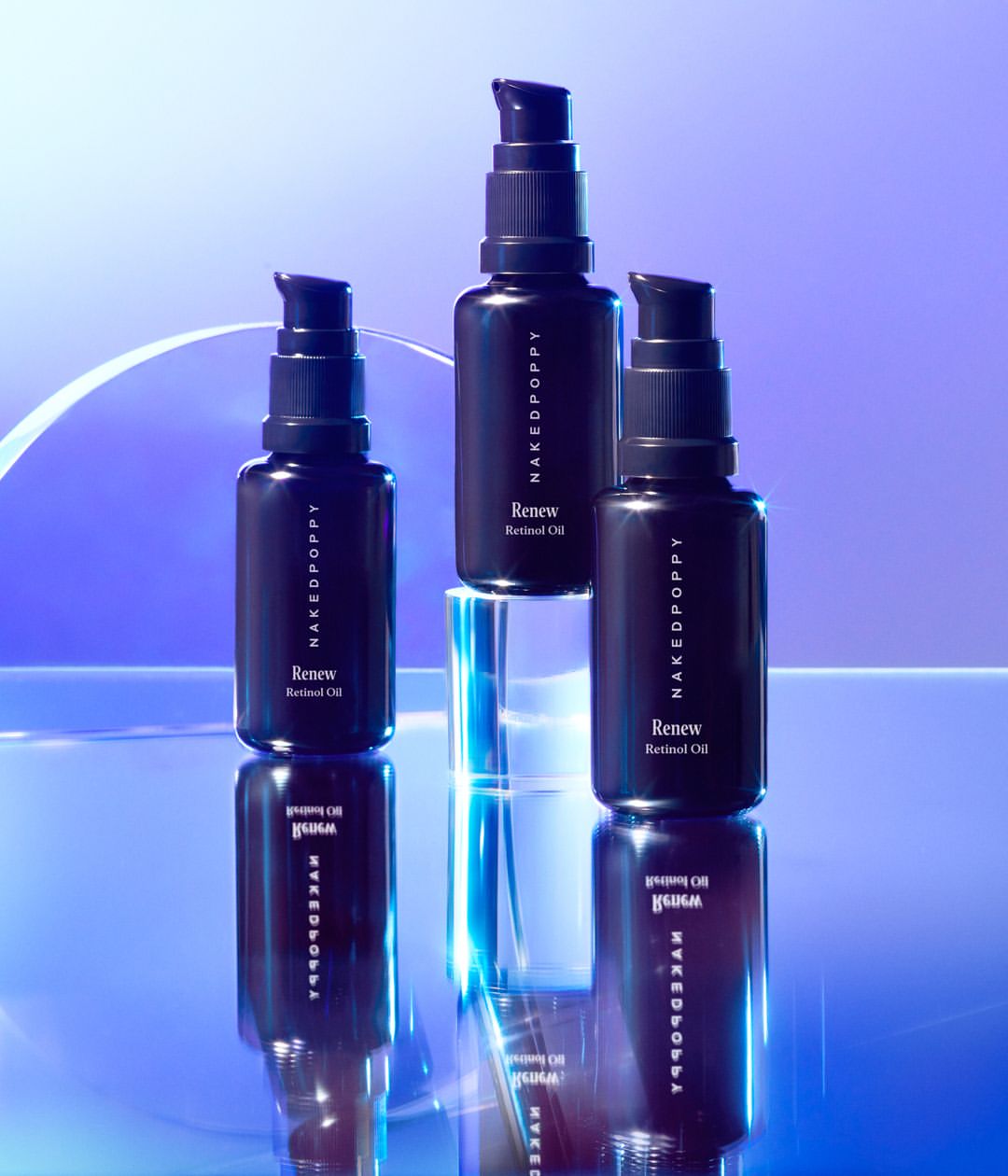 Three black bottles of Naked Poppy Renew Retinol Oil are displayed against a blue background, sitting on a reflective surface and clear block.