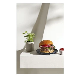 Gourmet burger on a dark plate with a small potted plant on a white ledge with harsh angled shadows.