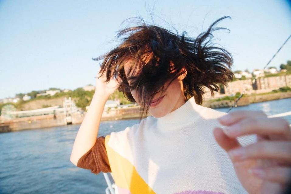 Young woman with short hair smiles on a boat against a backdrop of the coastline. The woman is wearing a patterned sweater.