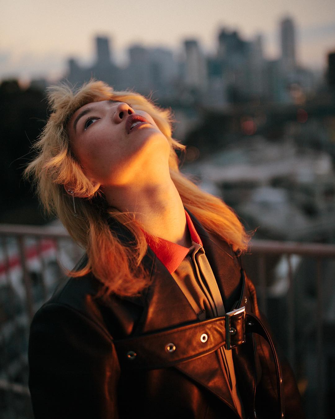 Woman with blonde hair and black jacket looks up at the city skyline during a sunset; warm lighting and urban background.