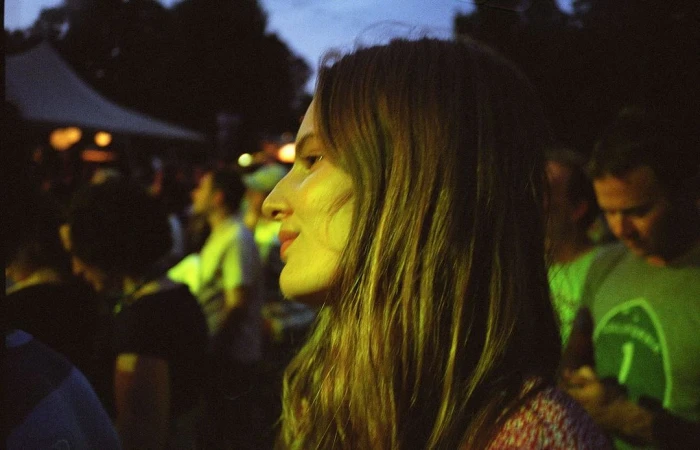 Profile portrait of an adult woman with long hair at an outdoor nighttime event, with a crowd and tent in the background.