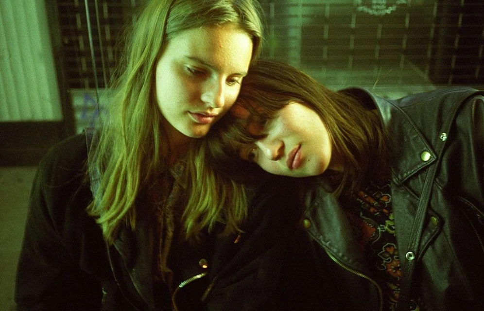 Two young women pose closely together, one with head resting on the other's shoulder, wearing leather jackets in a moody, green-toned indoor scene.