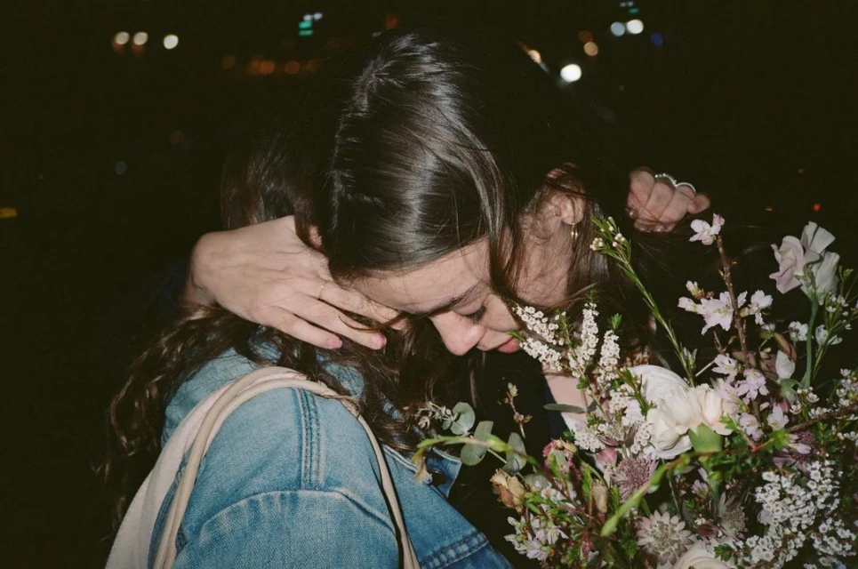 Close-up of two women embracing in dark surroundings, one carrying a large bouquet of white and green flowers in an intimate moment.