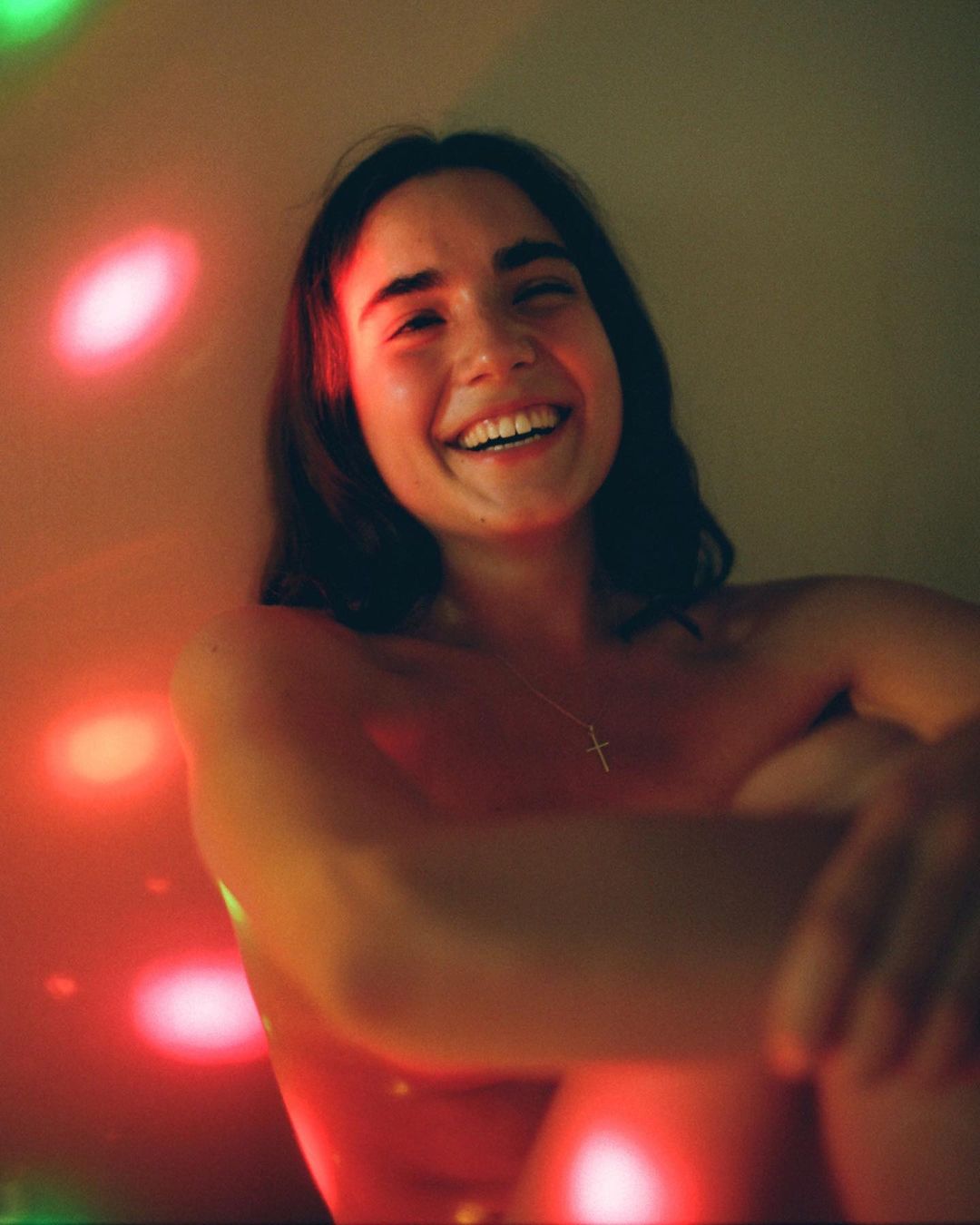 Close-up portrait of a smiling young adult woman with dark hair, fair skin, and a cross necklace, illuminated by red and green lights.