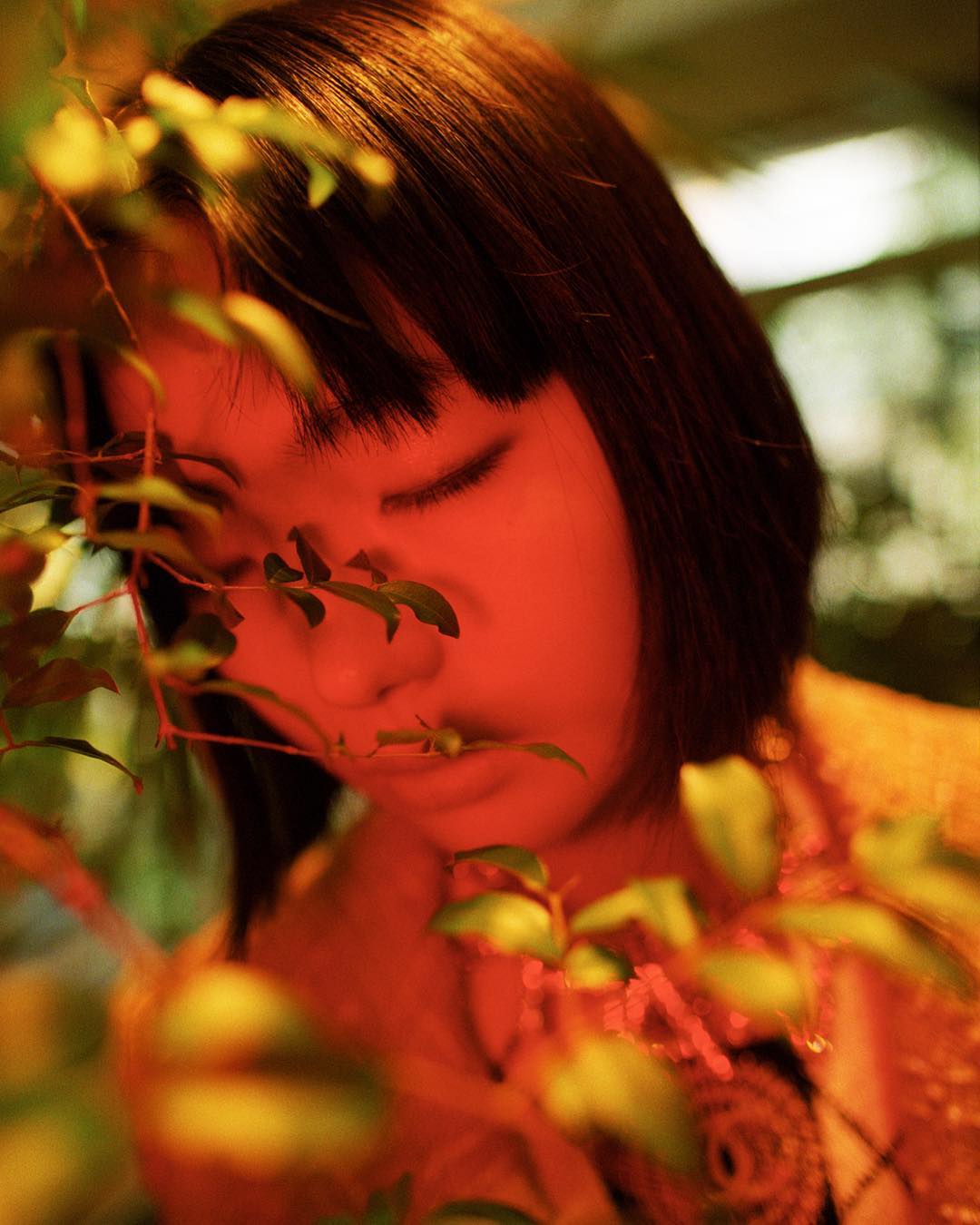Portrait of a young Asian woman with short black hair, eyes closed, framed by green leafy branches with a warm, golden color grade.