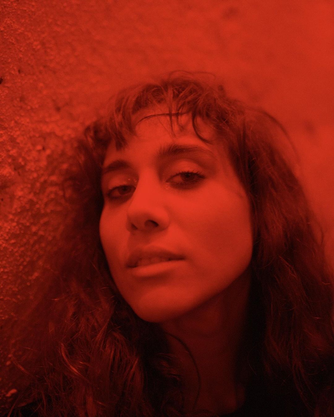 Portrait of a woman with long, wavy dark hair and light skin, illuminated by a red light, standing indoors.