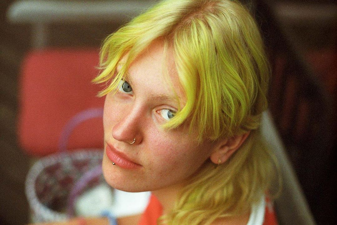 Close-up portrait of a young adult woman with brightly dyed yellow hair, fair skin, and blue eyes, looking directly at the camera.