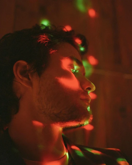 Close-up profile of an adult male with dark hair, beard and moustache, in vivid red and green light patterns against a dark backdrop.