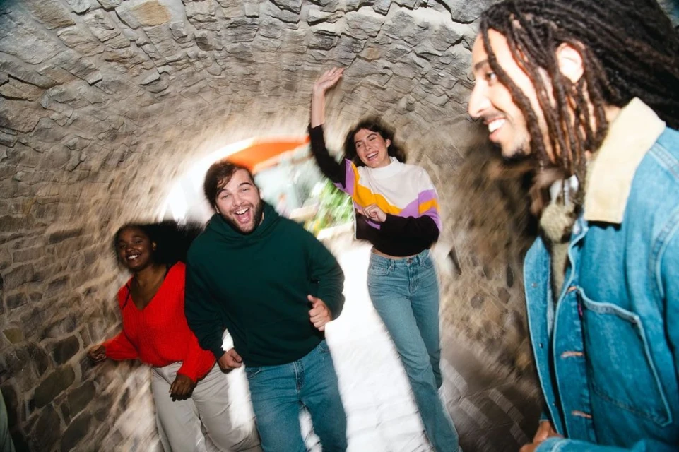 A group of four people in casual attire laughing and running through a stone tunnel. Three are in focus and one is slightly blurred.