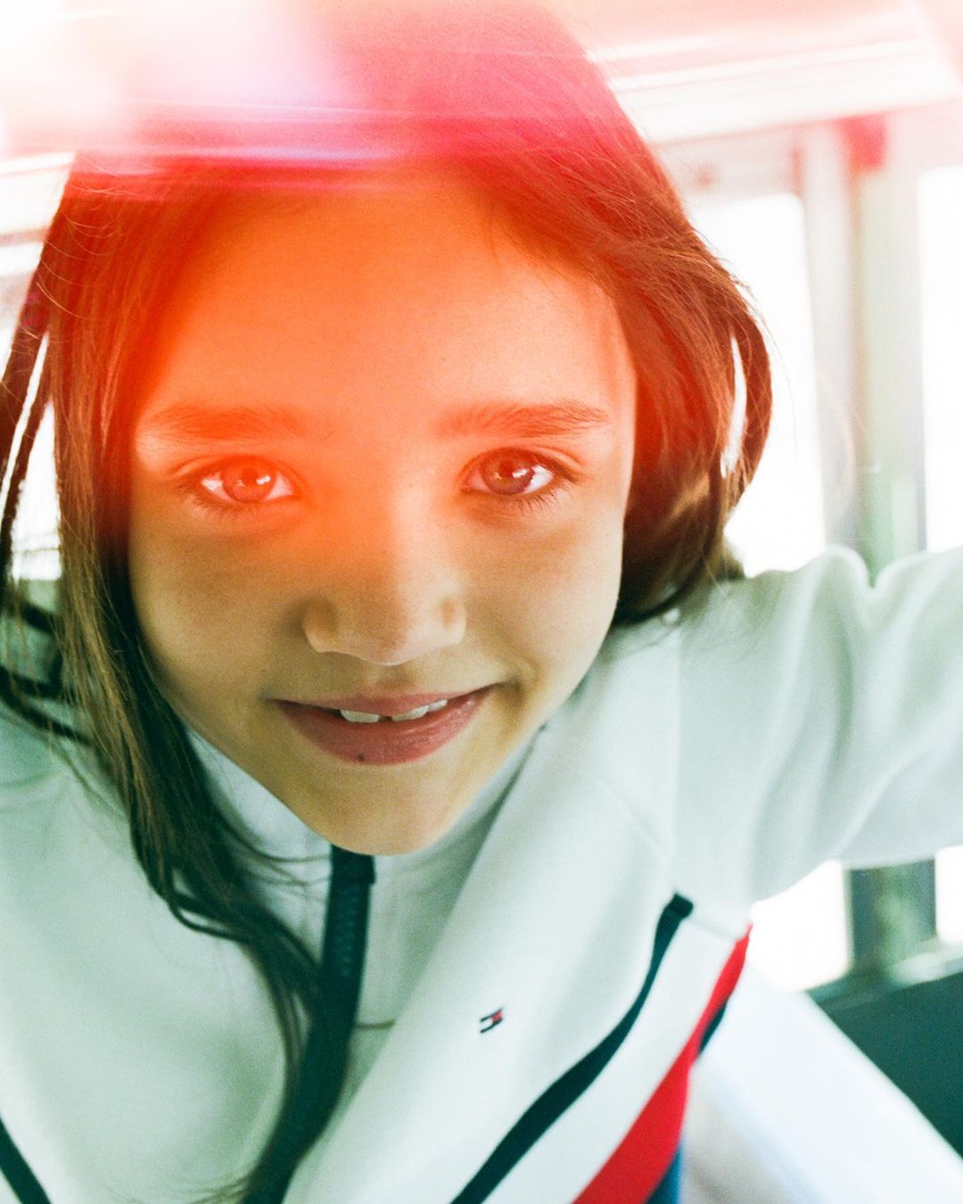 Close-up of a girl looking directly at the camera wearing a white athletic jacket with black trim indoors during the daytime.