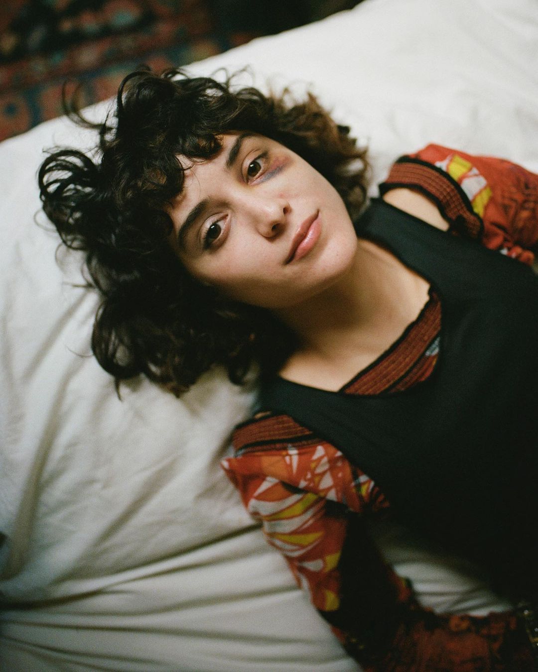 Indoor portrait of a young adult woman with curly brown hair lying on a white bed wearing a black dress over a patterned orange top.